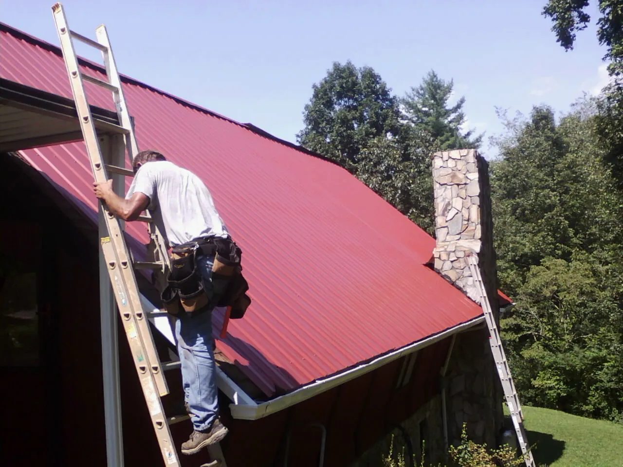 Roofer on a ladder beside a red metal roof and stone chimney — Soffit & Fascia Repair by Roofing Brothers in Udell